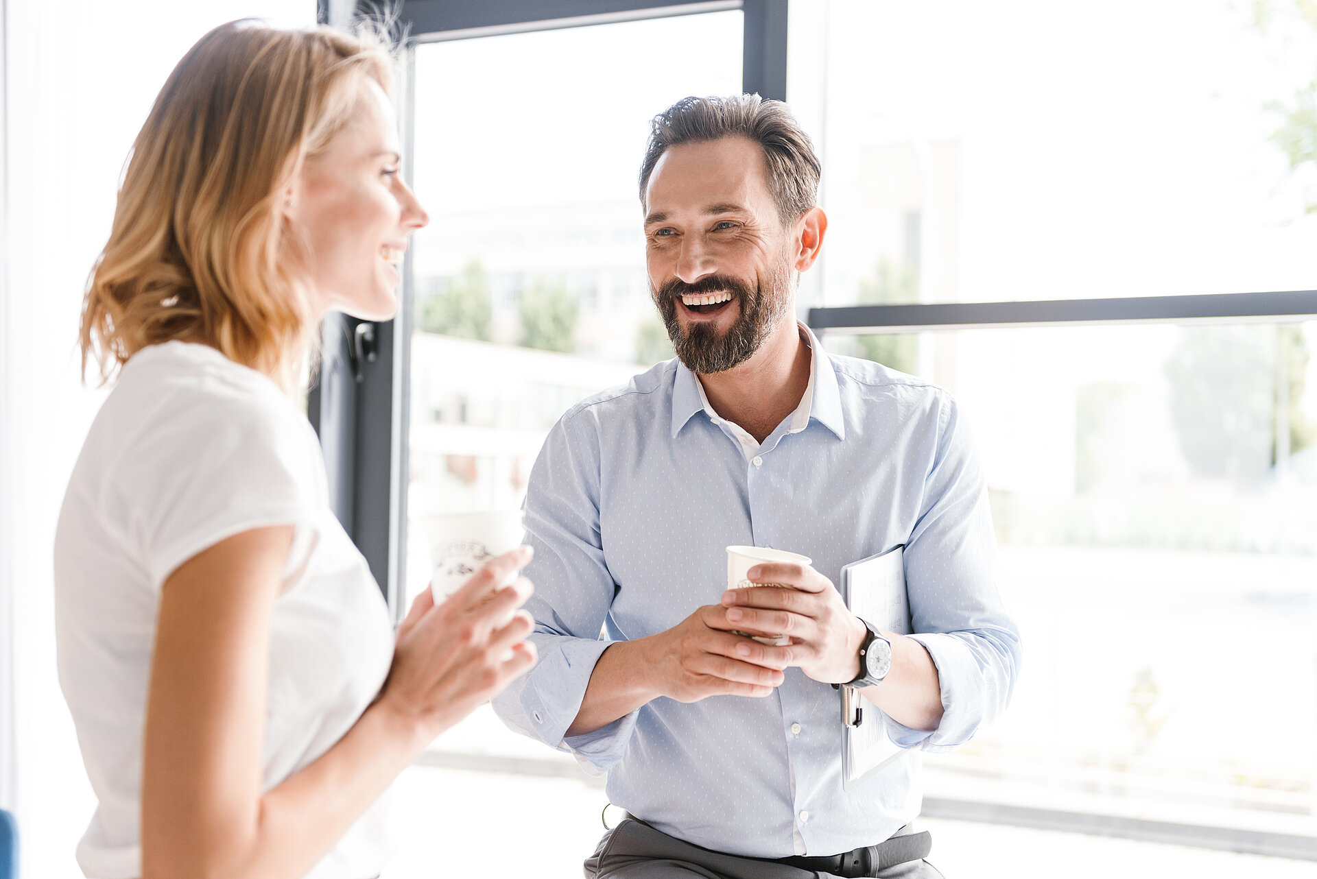 Mann und Frau stehen lachend in einem Büro, im Hintergrund eine Glasfront bei Sonnenschein