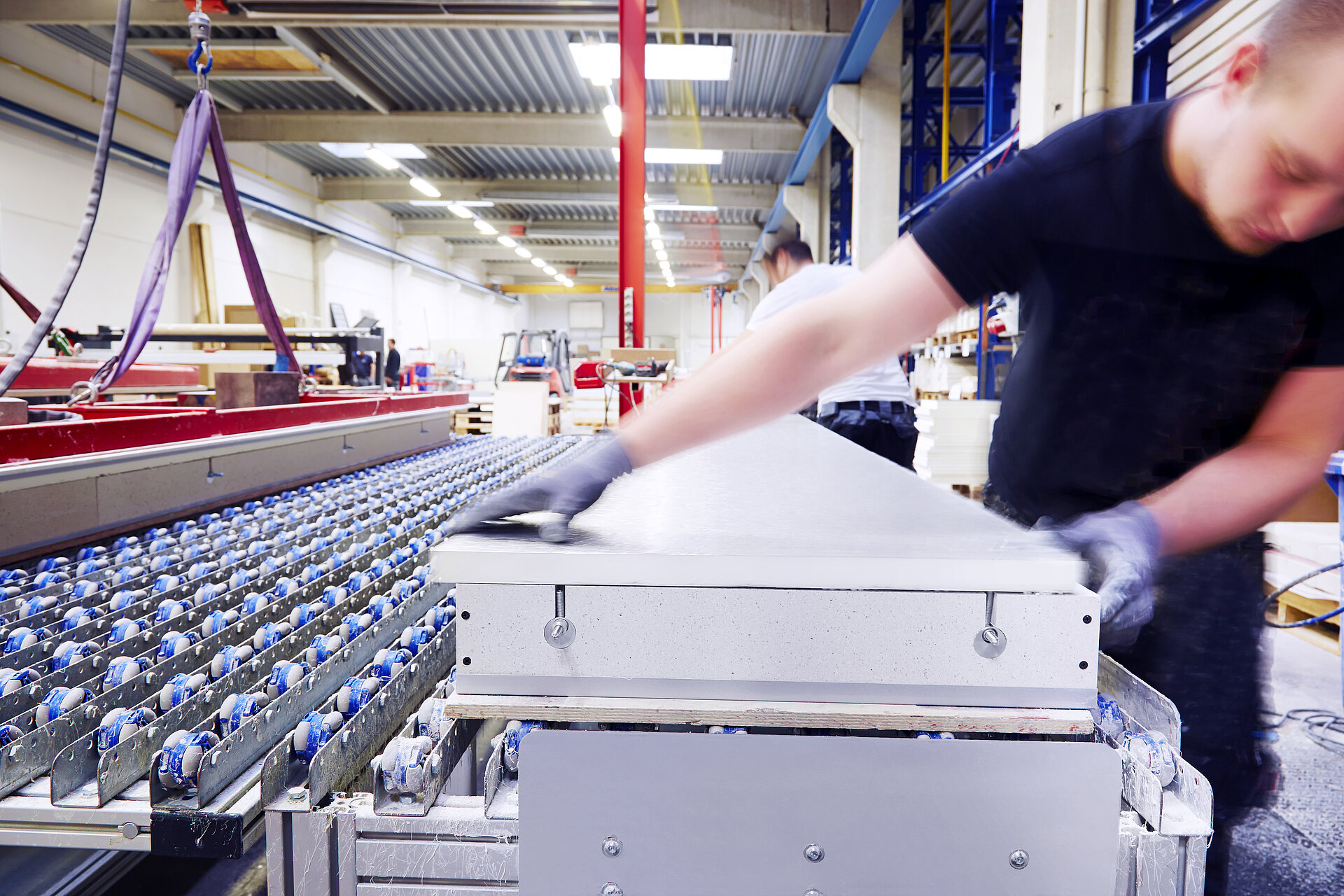 Mann blickt lachend auf ein Tablet und trägt einen Baustellenhelm in einem Lager. Im Hintergrund ein weiterer Mann