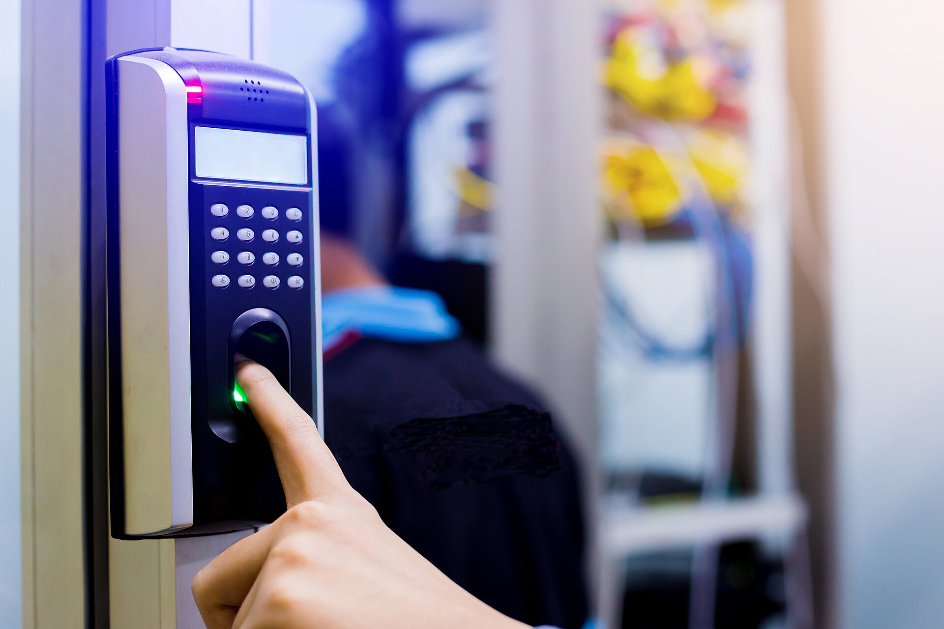 Sicherheitspad, welches von einem Finger bedient wird, dahinter ein Mann, der steht und an etwas arbeitet