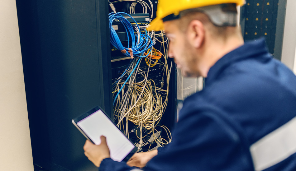 Mann mit blauer Arbeitskleidung und gelbem Helm steht vor einem Schrank mit vielen Kabeln und blickt dabei auf ein Tablet