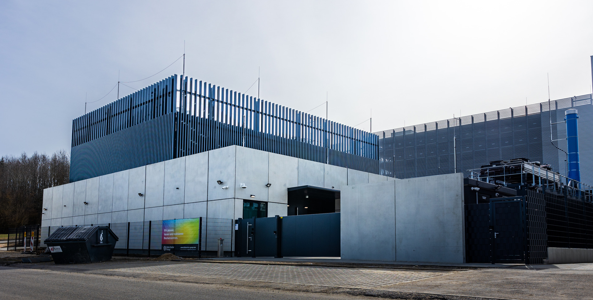 Rechenzentrum mit einer hellgrauen Fassade, rundum ein schwarzer Zaun aus Stahl und oben auf dem Dach Stahlstäbe was aussieht wie ein Gitter