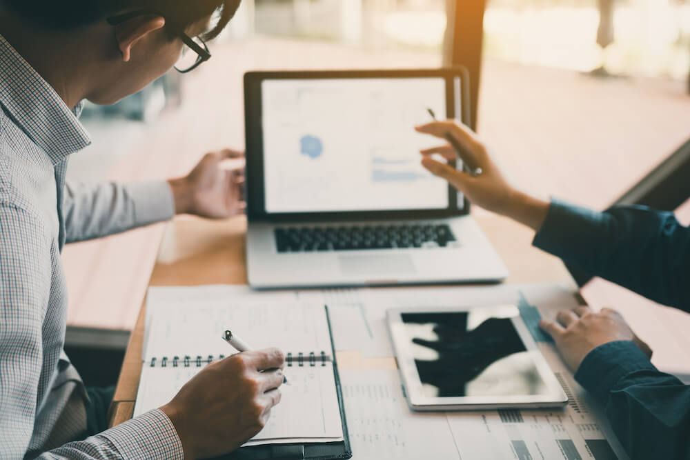 2 Personen, die auf einen Laptop schauen. Der eine notiert sich etwas auf einem Notizblock, der andere zeigt mit seinem Stift auf den BIldschirm