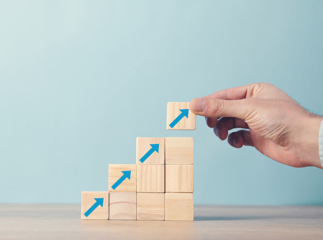 Mann, der Bauklötze aufeinander stapelt. erst 1 Klotz, dann 2, 3 und 4. Auf den jeweils oberen Klötzen ist ein Pfeil, der nach rechts oben zeigt, zu sehen.